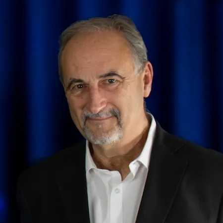 Older man with gray hair and beard wearing a dark suit and white shirt against a blue curtain background