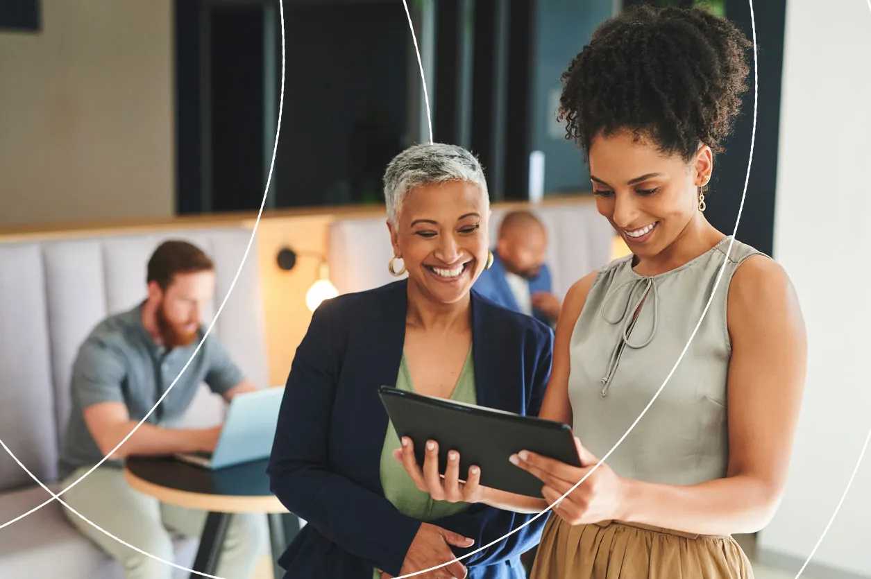 Two businesswomen standing together and smiling while looking at a tablet, with colleagues working in the background.