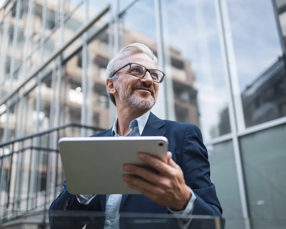 Smiling businessman in glasses using tablet device outside modern office building