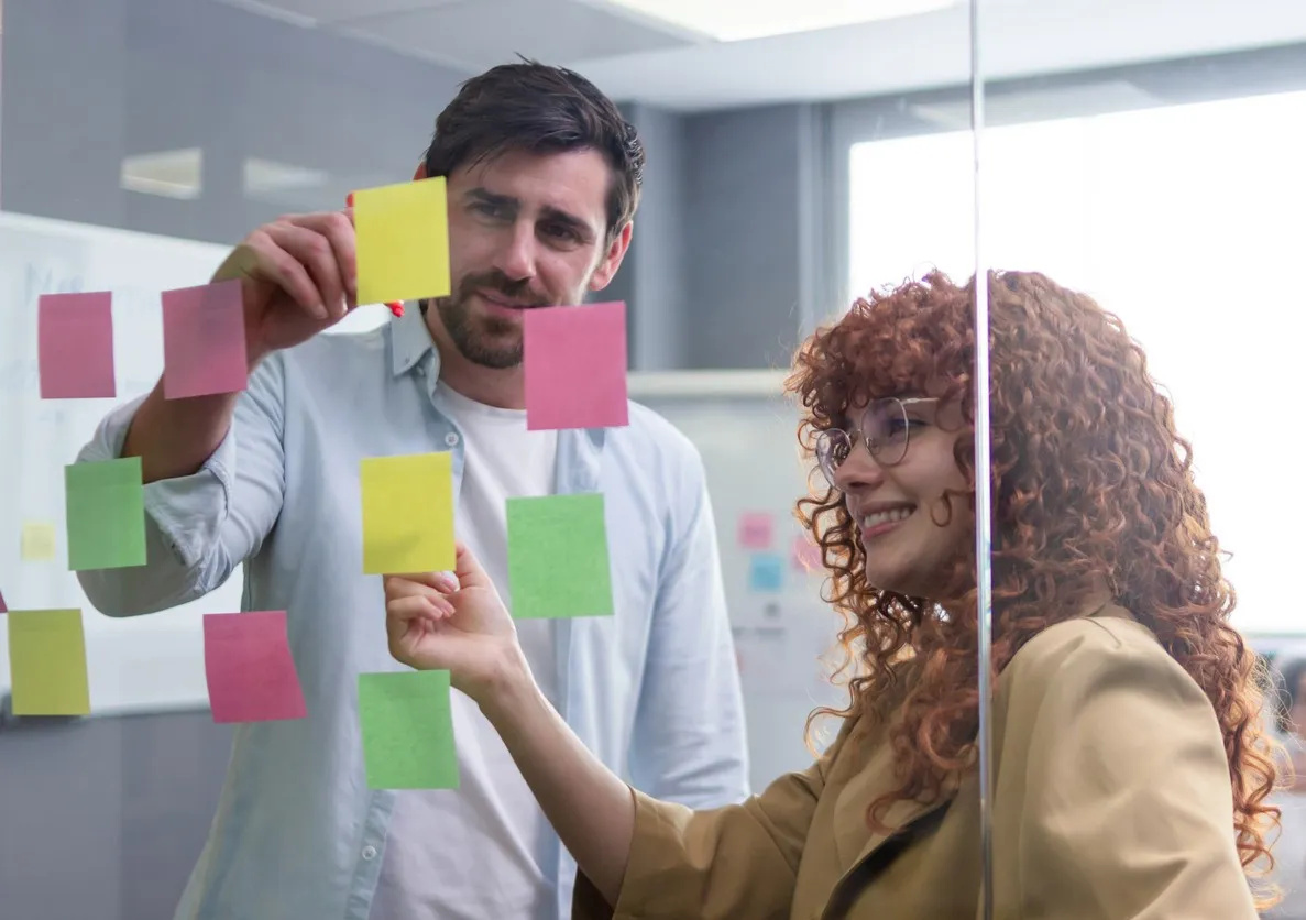 Two colleagues discussing colorful sticky notes on a glass wall in a modern office