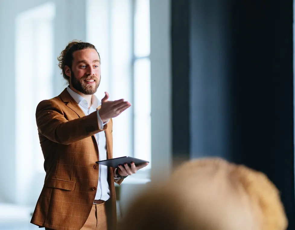 Man in brown jacket speaking and gesturing during a presentation