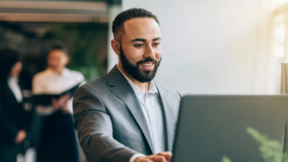 Professional in grey suit working on laptop at standing desk in bright office space
