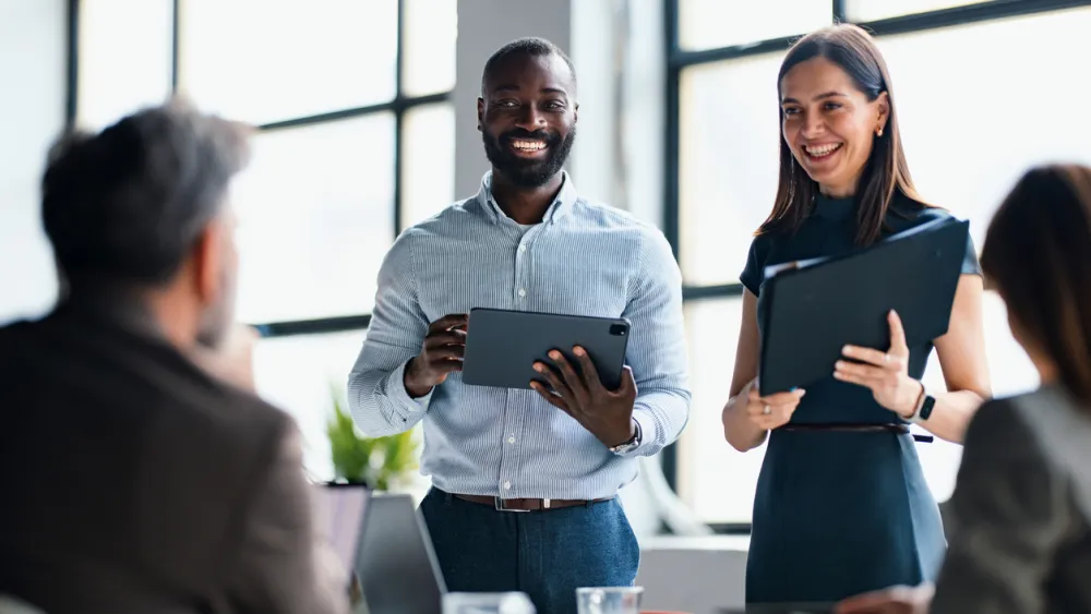 Two professionals presenting with tablets in a bright office to seated colleagues