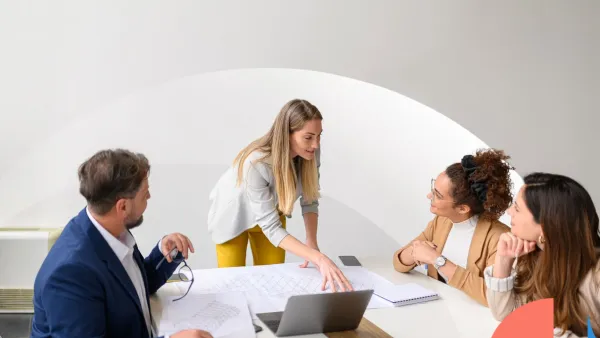 A woman presenting plans to three colleagues during a collaborative office meeting, with documents and a laptop on the table.