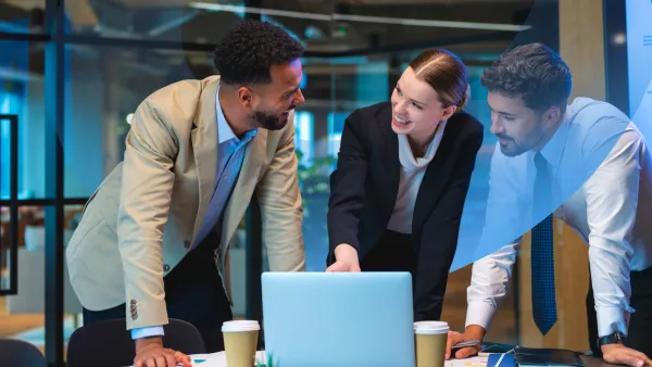 Two men and a woman in professional attire gathered around a table with a laptop, smiling and discussing a business plan.